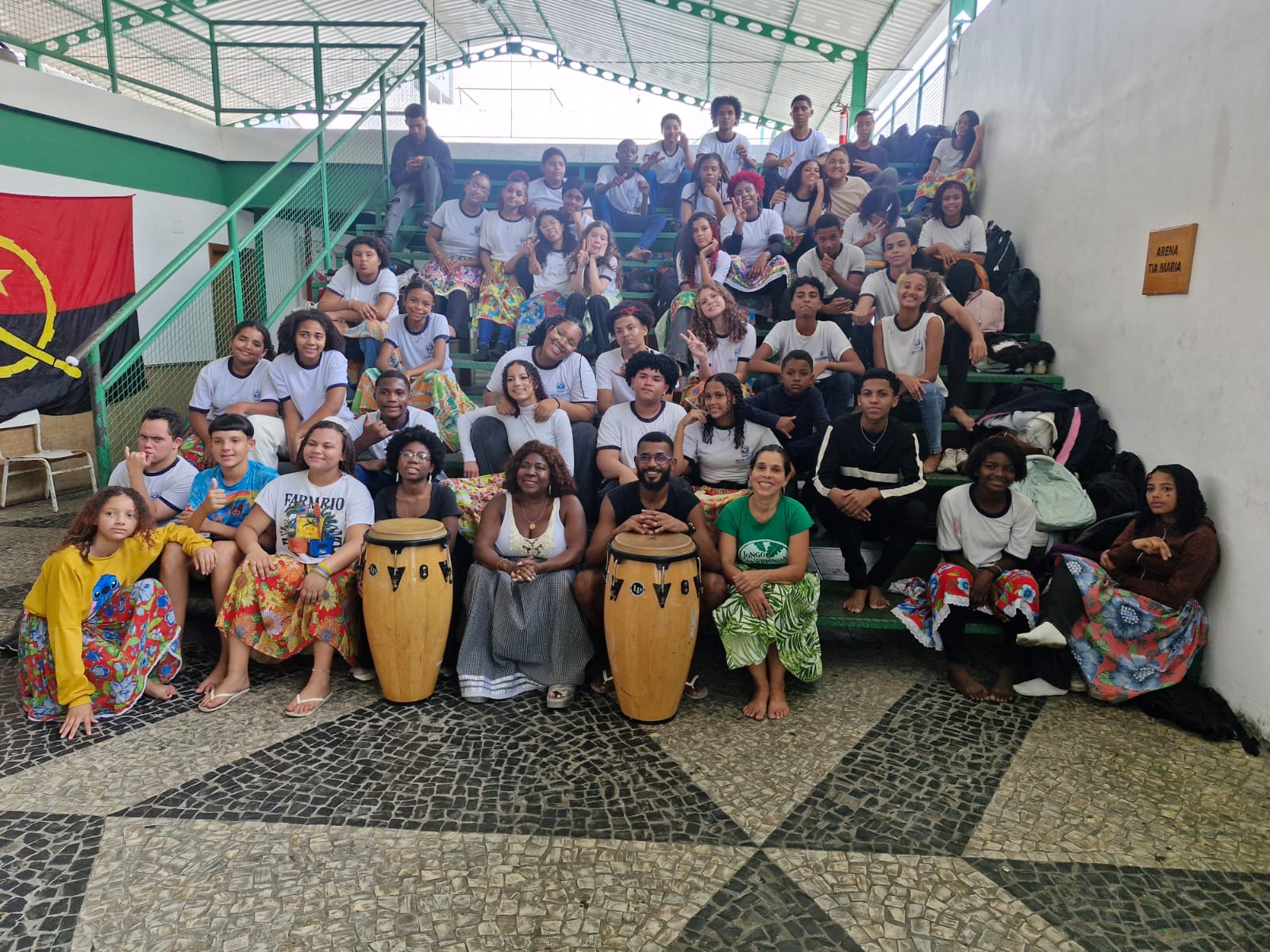Em foto desbotada, Tia Maria está em pé em uma varanda, à frente de uma porta marrom fechada. Ela é uma mulher negra e usa roupas brancas: vestido rendado com mangas curtas e lenço à cabeça, que cai acima de um ombro. Ela olha para frente e está com uma das mãos na cintura. Usa colar, pulseiras, relógio, anel, e sandálias com salto plataforma. A varanda tem azulejos à meia altura, brancos com estampas cinzas. Os patamares são brancos. Nela, há cadeiras com estofados brancos e vermelhos. No rodapé da foto, está escrito à caneta: 10-10-77.