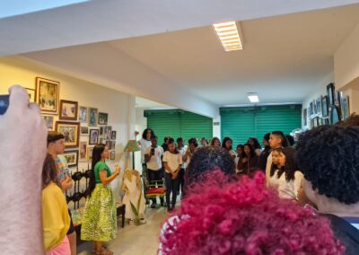 Uma mulher vestindo uma blusa verde e uma saia estampada conversa com um grupo de jovens em uma sala decorada com quadros nas paredes.