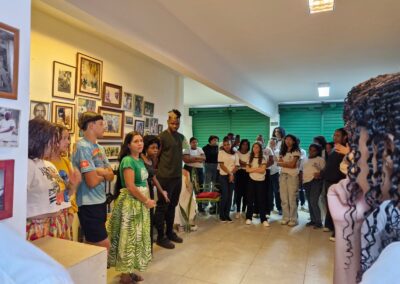 Um grupo de jovens se reuniu em uma sala com fotos na parede, ouvindo uma mulher falar.