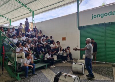 Um instrutor dá aula para um grupo de alunos sentados em arquibancadas em degraus, cada um segurando um tambor de percussão.