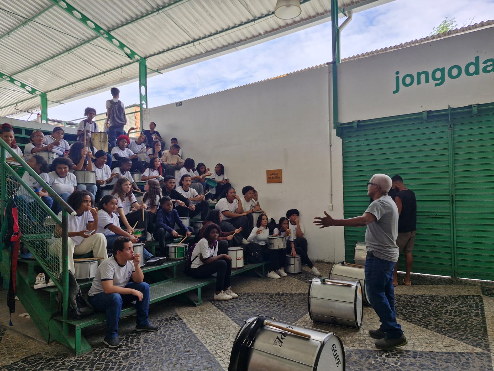 Um instrutor d&aacute; aula para um grupo de alunos sentados em arquibancadas em degraus, cada um segurando um tambor de percuss&atilde;o.