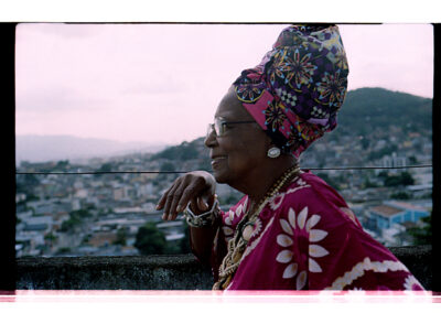 Uma mulher mais velha, com um lenço floral bem colorido na cabeça e uma blusa com estampa combinando, contemplando a cidade do alto de um telhado.