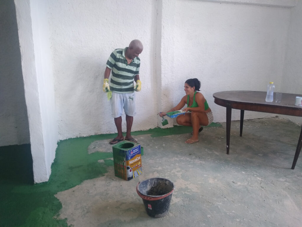 Um homem e uma mulher ajoelhados no chão, pintando a parte inferior de uma parede branca de verde.