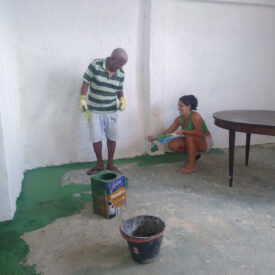 Um homem e uma mulher ajoelhados no chão, pintando a parte inferior de uma parede branca de verde.
