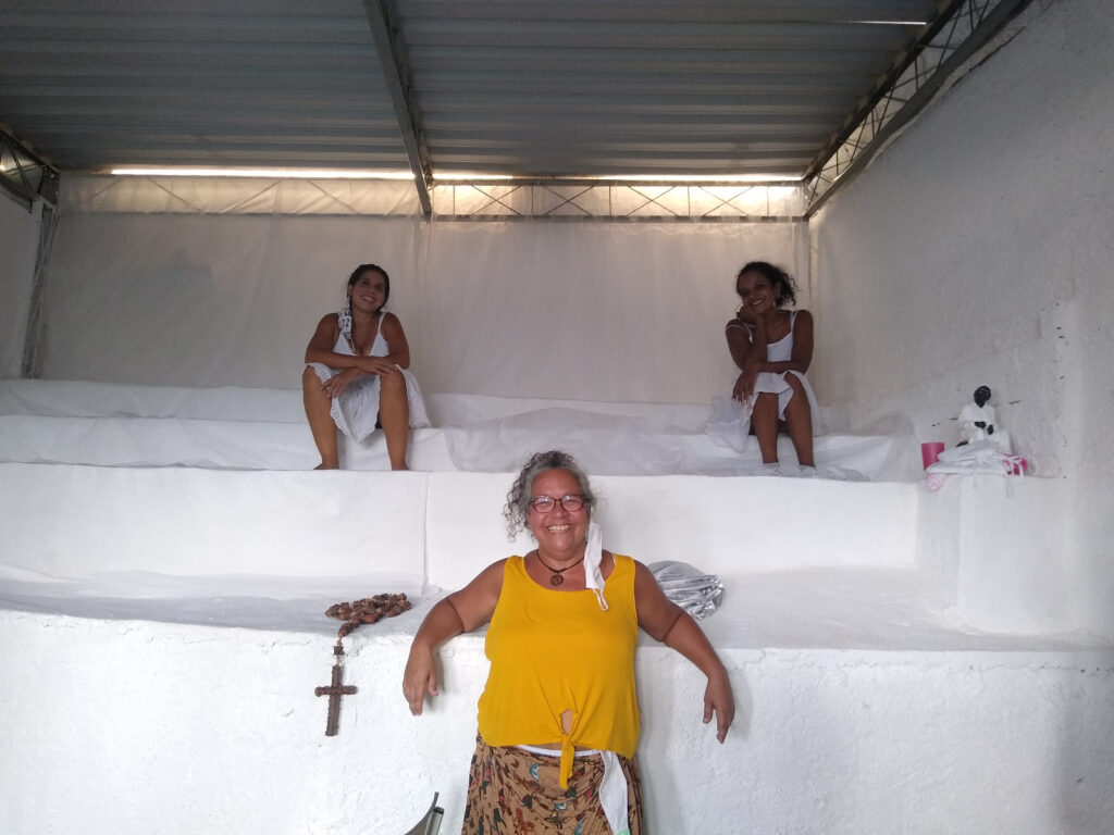 Três mulheres posando em uma sala com paredes brancas e vários níveis, sendo que duas estão sentadas nos níveis superiores e uma está em pé em primeiro plano, vestindo uma blusa amarela.
