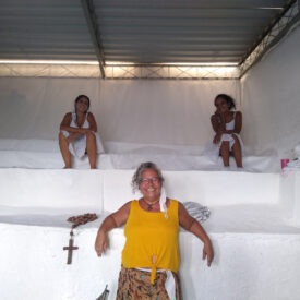 Três mulheres posando em uma sala com paredes brancas e vários níveis, sendo que duas estão sentadas nos níveis superiores e uma está em pé em primeiro plano, vestindo uma blusa amarela.
