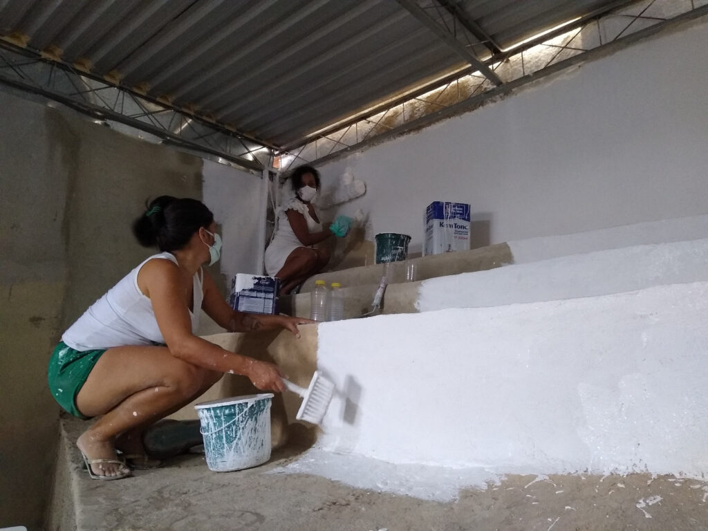 Duas mulheres usando máscaras faciais se agacham para pintar de branco uma escada de concreto.