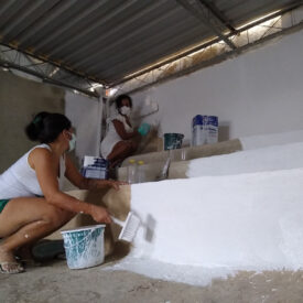 Duas mulheres usando máscaras faciais se agacham para pintar de branco uma escada de concreto.