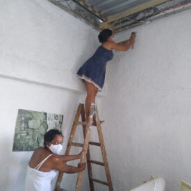 Uma mulher está em cima de uma escada de madeira, batendo com um martelo em uma parede branca, enquanto outra mulher, vestindo um vestido branco, está ao pé da escada.