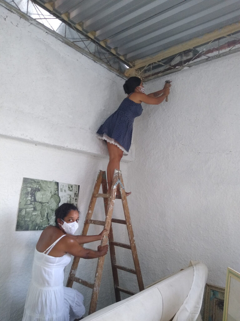 Uma mulher está em cima de uma escada de madeira, batendo com um martelo em uma parede branca, enquanto outra mulher, vestindo um vestido branco, está ao pé da escada.