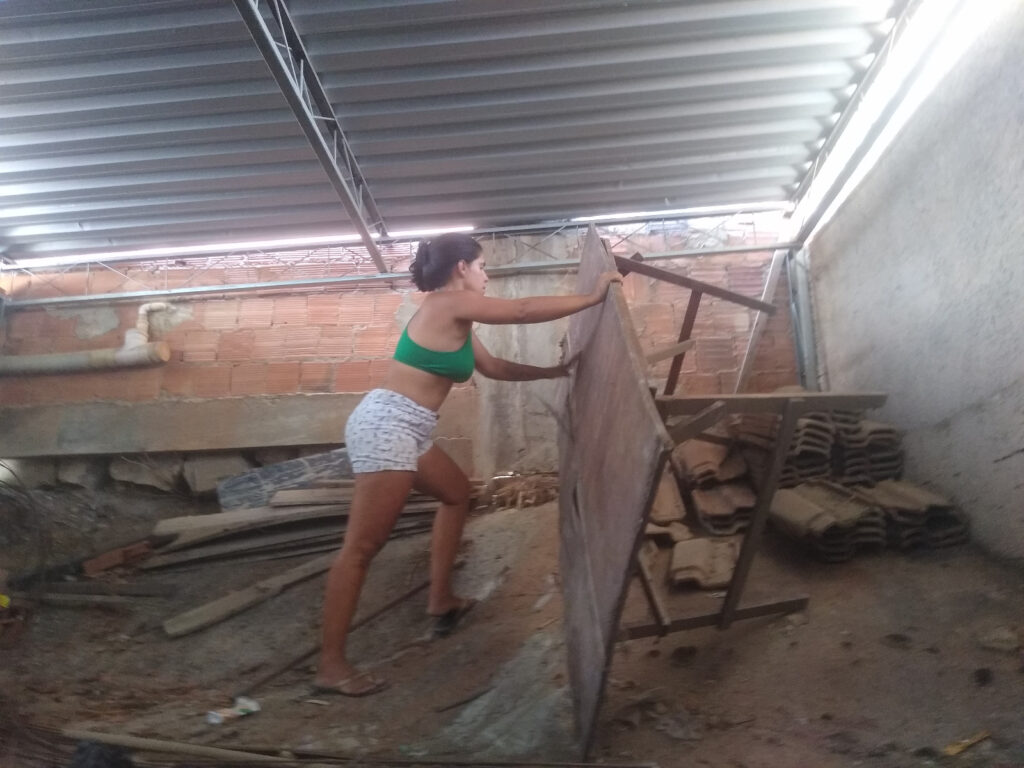Uma mulher vestindo um top curto verde e shorts estampados está encostada em uma grande placa de compensado em uma obra.