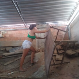 Uma mulher vestindo um top curto verde e shorts estampados está encostada em uma grande placa de compensado em uma obra.
