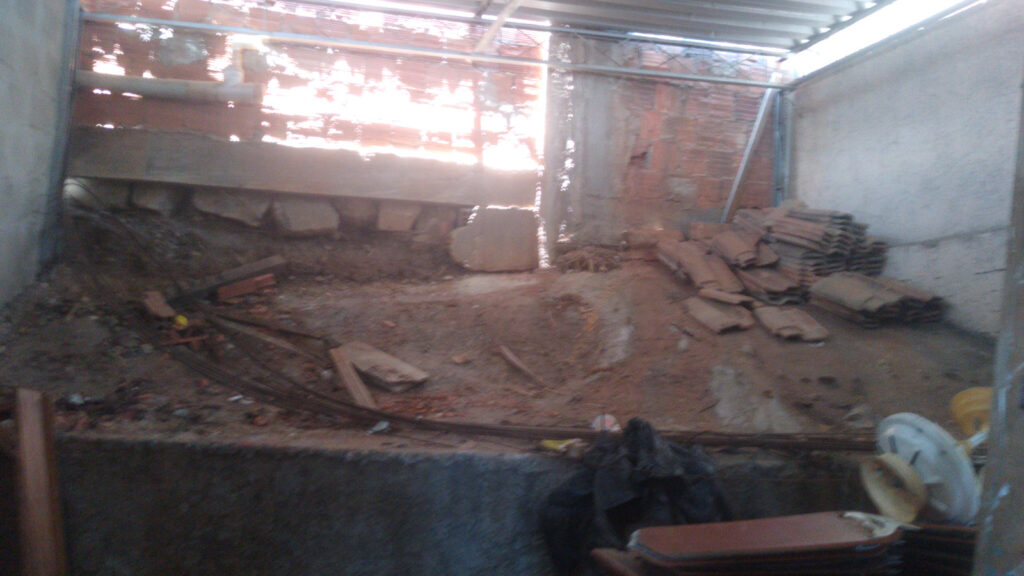 Um canteiro de obras com piso de terra batida, telhas empilhadas e uma parede aberta voltada para uma luz intensa.