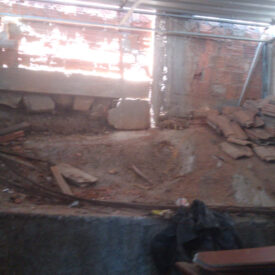Um canteiro de obras com piso de terra batida, telhas empilhadas e uma parede aberta voltada para uma luz intensa.