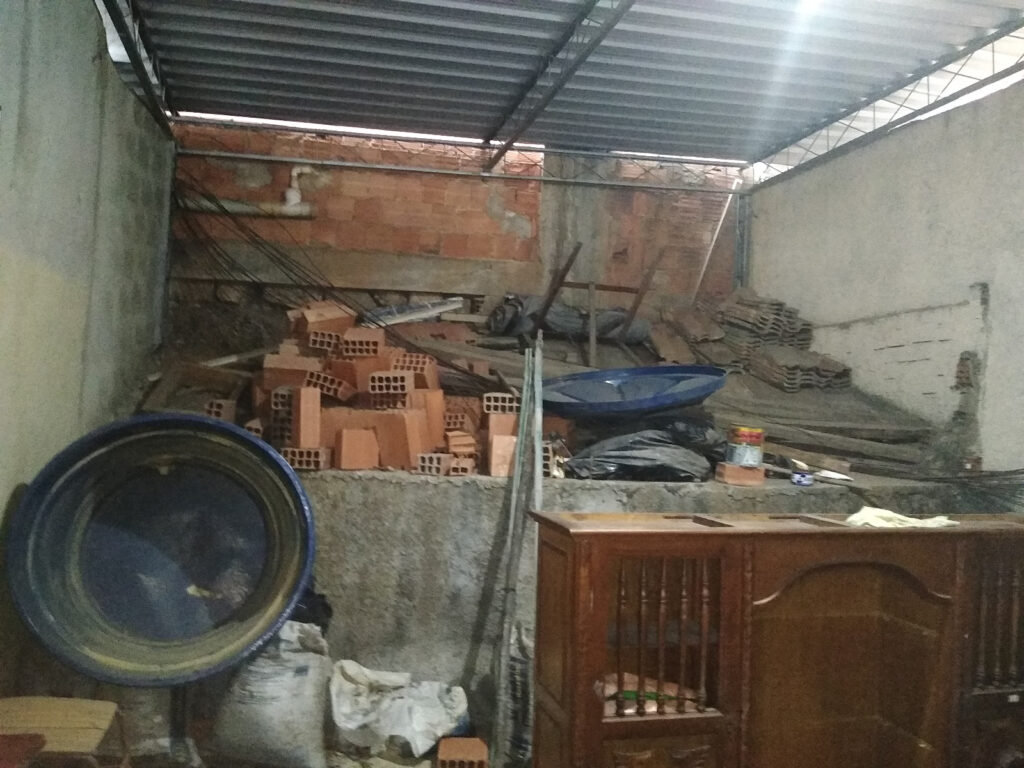 Um cômodo inacabado com uma pilha de tijolos vermelhos, uma bacia de plástico azul e um armário de madeira sob um telhado de metal.