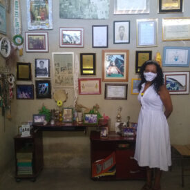 Uma mulher vestida com um vestido branco e usando máscara está em uma sala cujas paredes estão cobertas de fotografias emolduradas, diplomas e prêmios.