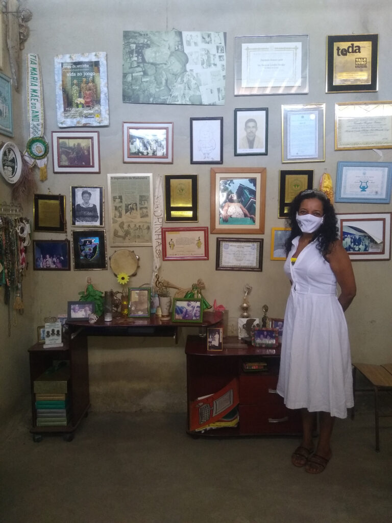 Uma mulher vestida com um vestido branco e usando máscara está em uma sala cujas paredes estão cobertas de fotografias emolduradas, diplomas e prêmios.