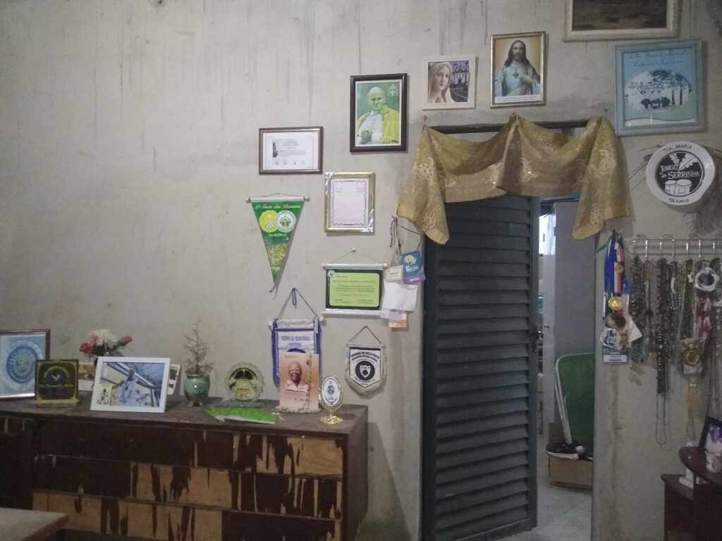Parede decorada com várias imagens religiosas emolduradas, bandeirinhas, certificados e uma coleção de colares pendurados.