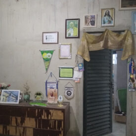 Parede decorada com várias imagens religiosas emolduradas, bandeirinhas, certificados e uma coleção de colares pendurados.