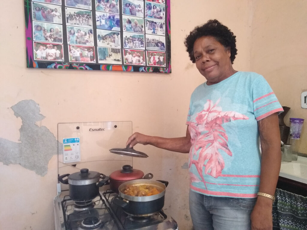 Uma mulher está em frente ao fogão, levantando a tampa de uma panela com comida.