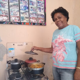 Uma mulher está em frente ao fogão, levantando a tampa de uma panela com comida.