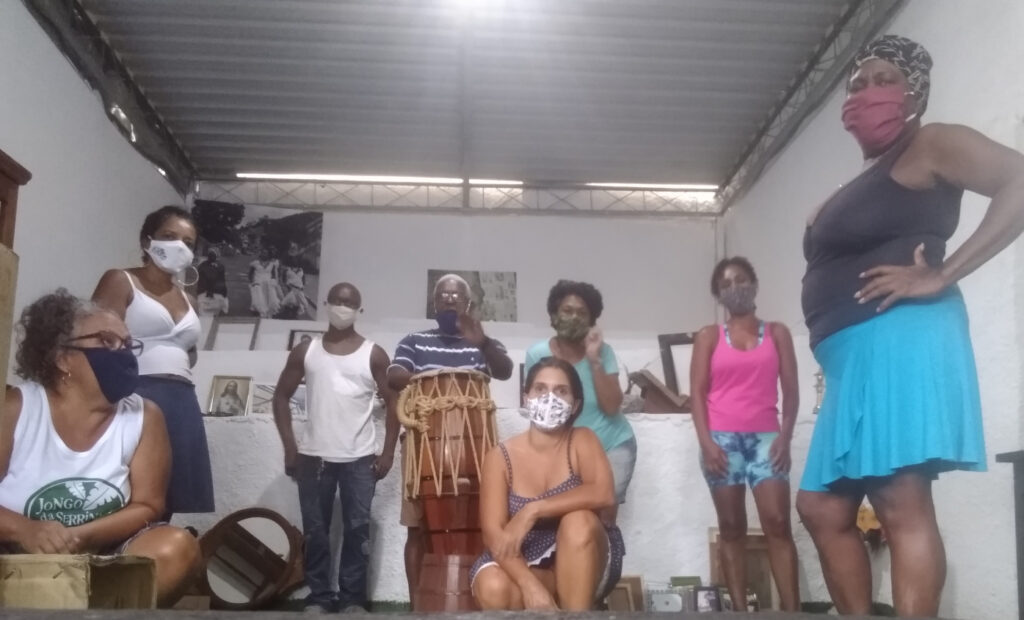 Oito pessoas de várias idades, em pé e ajoelhadas em uma sala, todas usando máscaras faciais, com um homem em pé atrás de um grande tambor de madeira.