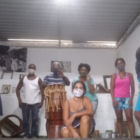 Oito pessoas de várias idades, em pé e ajoelhadas em uma sala, todas usando máscaras faciais, com um homem em pé atrás de um grande tambor de madeira.